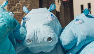 Three people wearing large, blue hippo costumes stand close together outdoors, with buildings and a stone wall in the background. The costumes feature exaggerated hippo faces and nostrils.