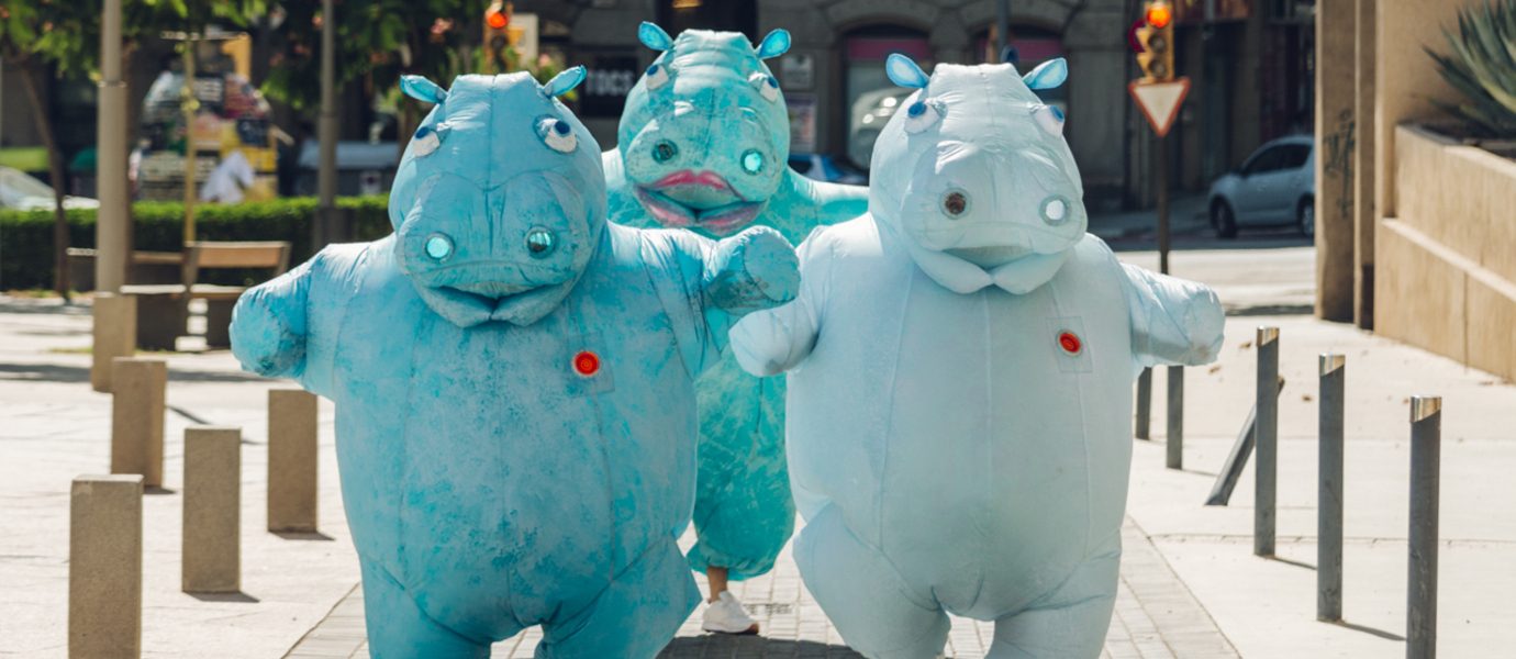 Three people dressed in large, blue hippopotamus costumes walk energetically down a sunny city street, surrounded by trees, buildings, and street signs.