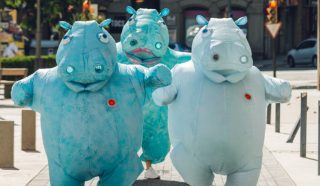 Three people dressed in large, blue hippopotamus costumes walk energetically down a sunny city street, surrounded by trees, buildings, and street signs.