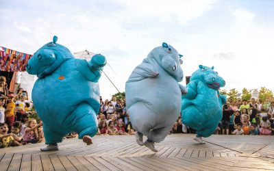 Three people in large, blue inflatable hippo costumes dance energetically on an outdoor wooden stage, with a cheering crowd and colourful decorations in the background.