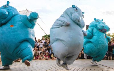 Three people in inflatable blue hippo costumes dance energetically on an outdoor stage, whilst a large crowd of children and adults watch and smile in the background.