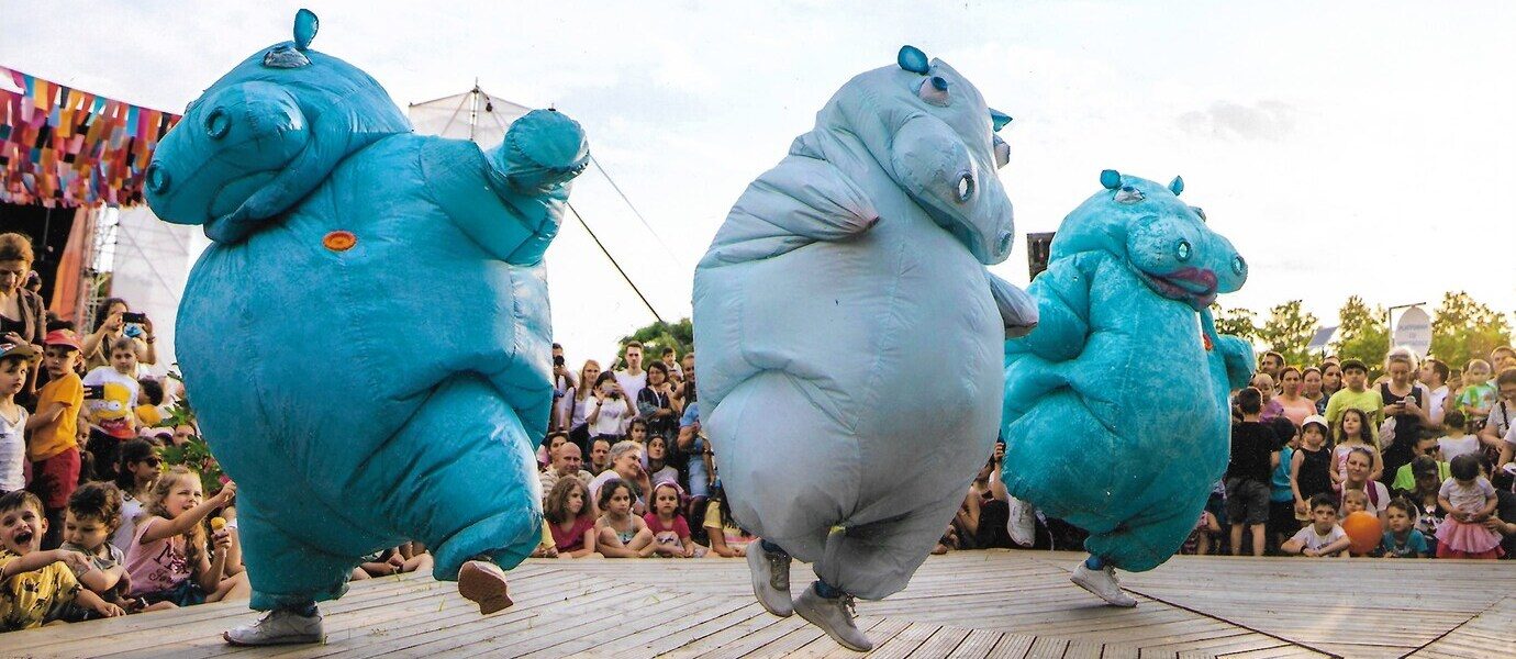 Three performers in large, blue inflatable hippo costumes dance energetically on a wooden stage, entertaining a crowd of people, including many children, seated and standing in the background outdoors.