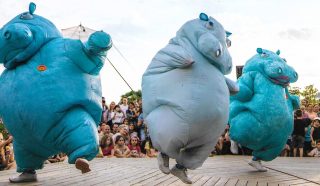 Three performers in large, blue inflatable hippo costumes dance energetically on a wooden stage, entertaining a crowd of people, including many children, seated and standing in the background outdoors.