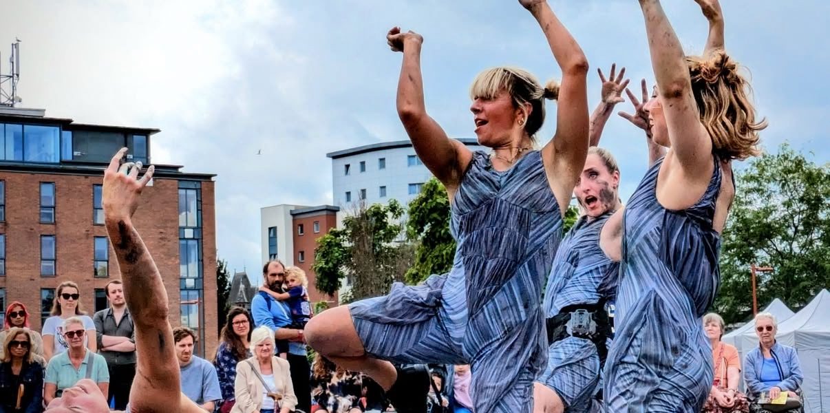 Four people in matching blue patterned outfits perform an expressive dance outdoors, with dramatic poses and gestures, as an audience watches in the background. Modern buildings and cloudy skies are visible.