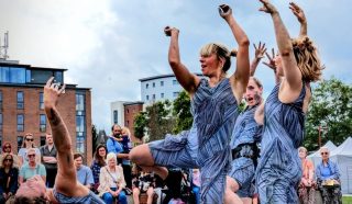 Four people in matching blue patterned outfits perform an expressive dance outdoors, with dramatic poses and gestures, as an audience watches in the background. Modern buildings and cloudy skies are visible.