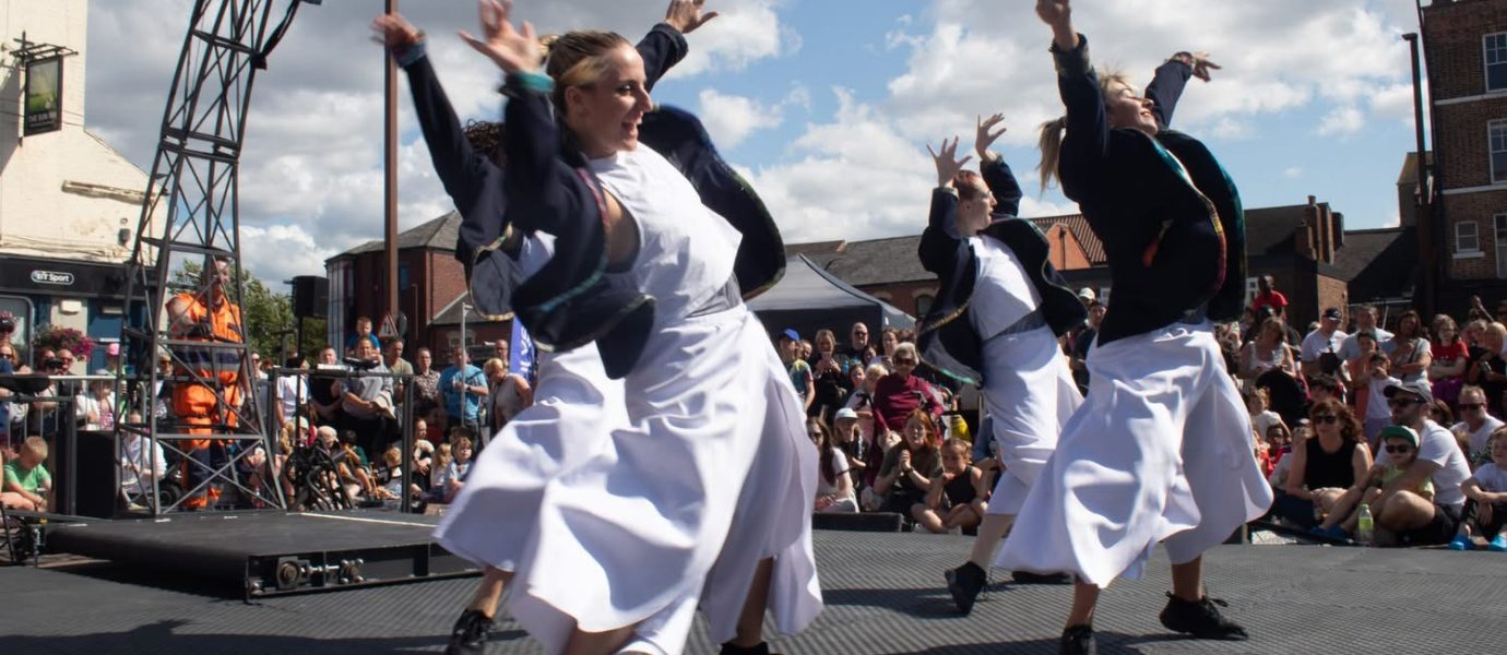 Four dancers wearing white dresses and dark jackets perform energetically on an outdoor stage, raising their arms. A crowd watches in the background under a partly cloudy sky.