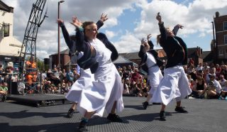 Four dancers wearing white dresses and dark jackets perform energetically on an outdoor stage, raising their arms. A crowd watches in the background under a partly cloudy sky.