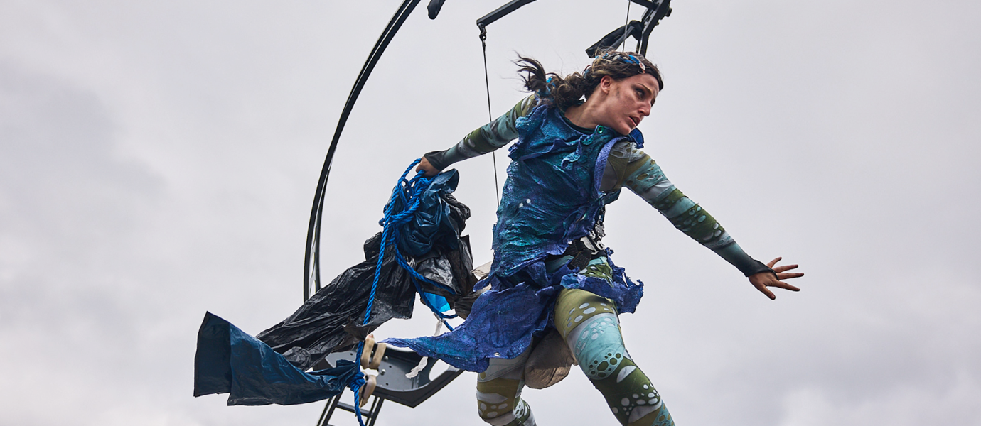 A performer in a blue and green costume is suspended in mid-air, holding flowing fabric, with one arm outstretched. A metal structure and ladder are behind her against a cloudy sky.