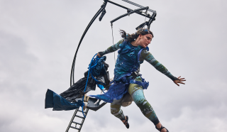 A performer in a blue and green costume is suspended in mid-air, holding flowing fabric, with one arm outstretched. A metal structure and ladder are behind her against a cloudy sky.