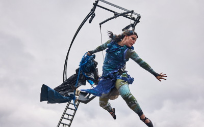 A performer in a blue and green costume is suspended in mid-air, holding flowing fabric, with one arm outstretched. A metal structure and ladder are behind her against a cloudy sky.