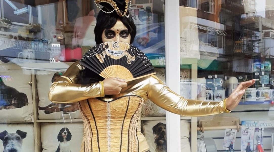 A person in gold clothing and skeletal face paint holds a black fan in front of their face. They are standing outside a shop with dog-themed decor visible inside through the window.