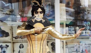 A person in gold clothing and skeletal face paint holds a black fan in front of their face. They are standing outside a shop with dog-themed decor visible inside through the window.