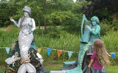 Two people dressed as mermaids pose outdoors; one in silver with a net and the other in green playing a harp. A child with long hair stands in front, and colourful bunting hangs in the background amidst trees and greenery.