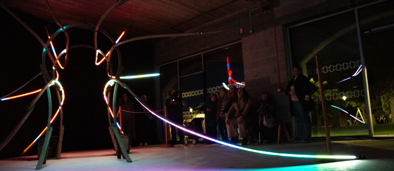 Colourful neon light installation with curved tubes glows in a dark industrial space. People stand and sit along the wall, observing the artwork. Reflections from the lights appear on the floor and nearby window.