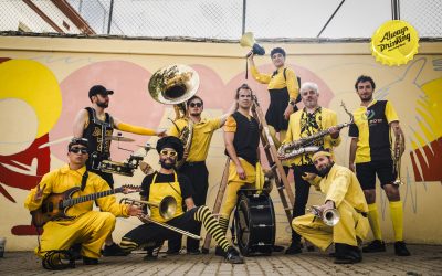 Nine musicians in yellow and black outfits pose with brass, guitar, and percussion instruments in front of a colourful wall mural. A yellow circular sign in the background reads Always Drinking.