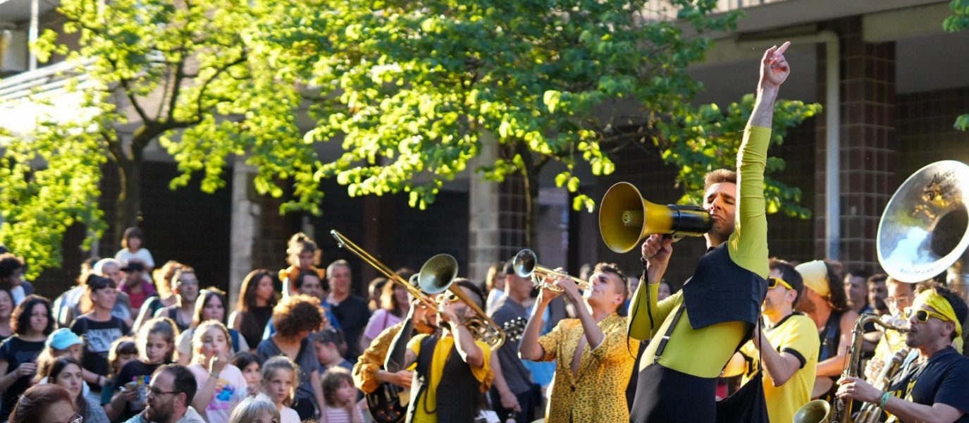 A lively brass band in yellow costumes performs energetically outdoors for a seated crowd, with one performer raising an arm. Trees and a building are in the background; a yellow “Always Printing” sign is visible above.