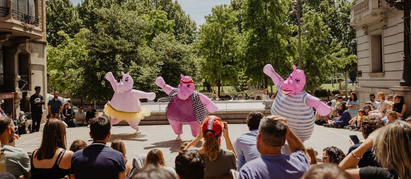 Three people in large, pink hippopotamus costumes perform for a seated outdoor audience on steps, with trees and historic buildings in the background on a sunny day.