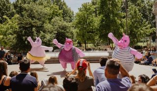 Three people in large, pink hippopotamus costumes perform for a seated outdoor audience on steps, with trees and historic buildings in the background on a sunny day.