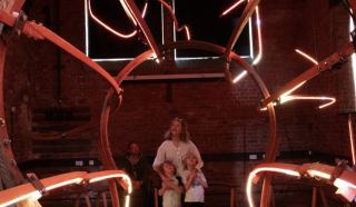 A woman and two children stand inside a large circular art installation with glowing red-orange neon lights in a dimly lit room with brick walls. An adult is seated in the background.