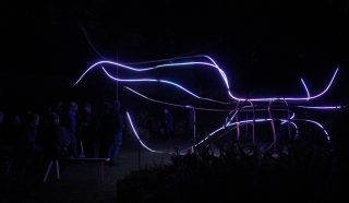 A group of people observes a glowing outdoor light installation at night, featuring curving lines of purple and blue neon lights arranged in an abstract shape. The surroundings are dimly lit with silhouettes visible.