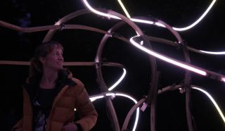 A woman in a yellow jacket stands at night, looking to the side and smiling, surrounded by curved wooden structures illuminated by glowing, looping neon lights.
