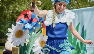A woman in a blue costume with a cloudy collar and hat entertains children outdoors, holding a tennis racquet decorated with bees and flowers. Large artificial flowers and greenery are in the background.