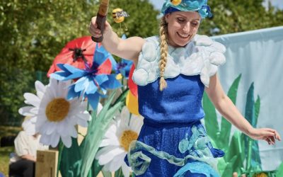 A woman in a blue costume with ocean-themed decorations holds a netted racquet and performs outdoors for children in blue hats, with large artificial flowers and greenery in the background.