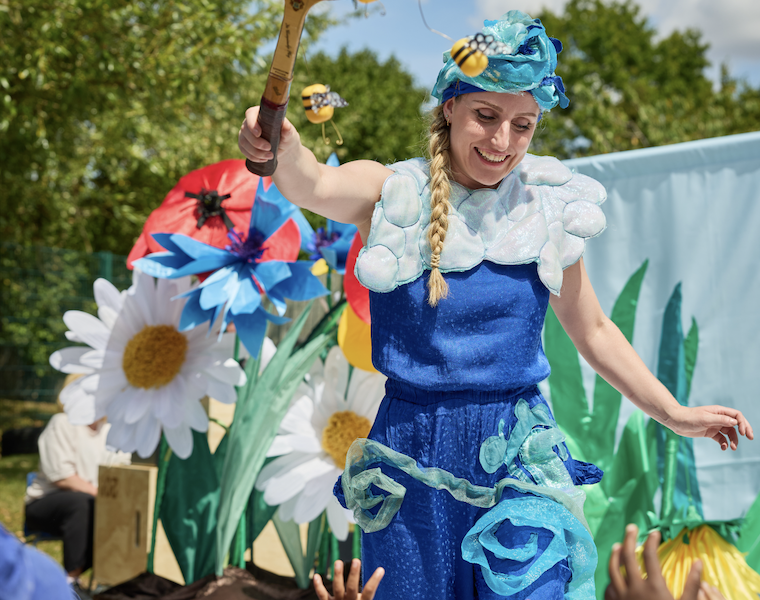 A woman in a blue costume with a cloudy collar and hat entertains children outdoors, holding a tennis racquet decorated with bees and flowers. Large artificial flowers and greenery are in the background.