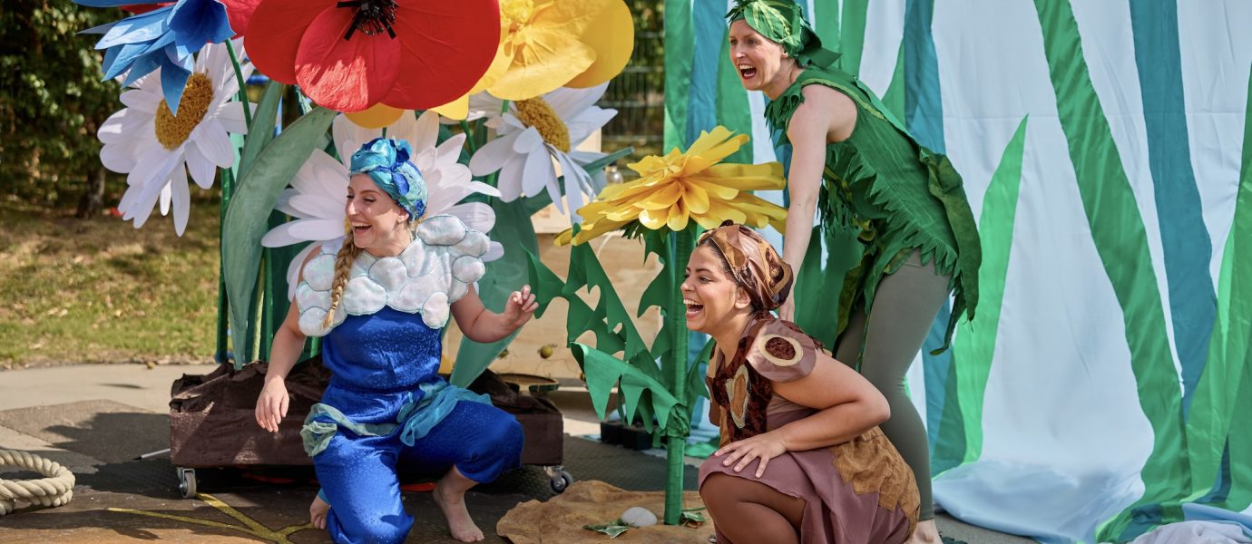 Three people dressed in colourful flower and plant costumes perform outdoors beside large, artificial flowers and a green backdrop, laughing and interacting with each other enthusiastically.