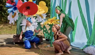 Three people dressed in colourful flower and plant costumes perform outdoors beside large, artificial flowers and a green backdrop, laughing and interacting with each other enthusiastically.