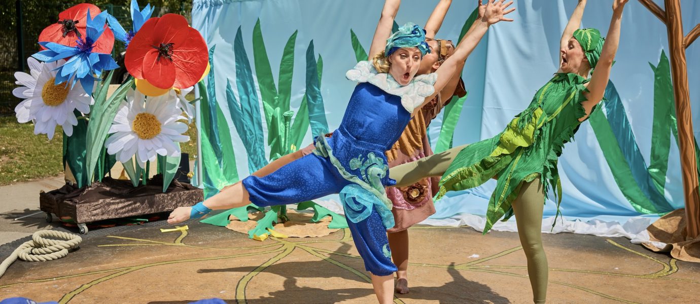 Three performers in colourful costumes strike dynamic poses during an outdoor play with a nature-themed backdrop, while children in blue hats watch from the audience. Large flowers and green plants decorate the stage.