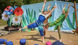 Three performers in colourful costumes strike dynamic poses during an outdoor play with a nature-themed backdrop, while children in blue hats watch from the audience. Large flowers and green plants decorate the stage.