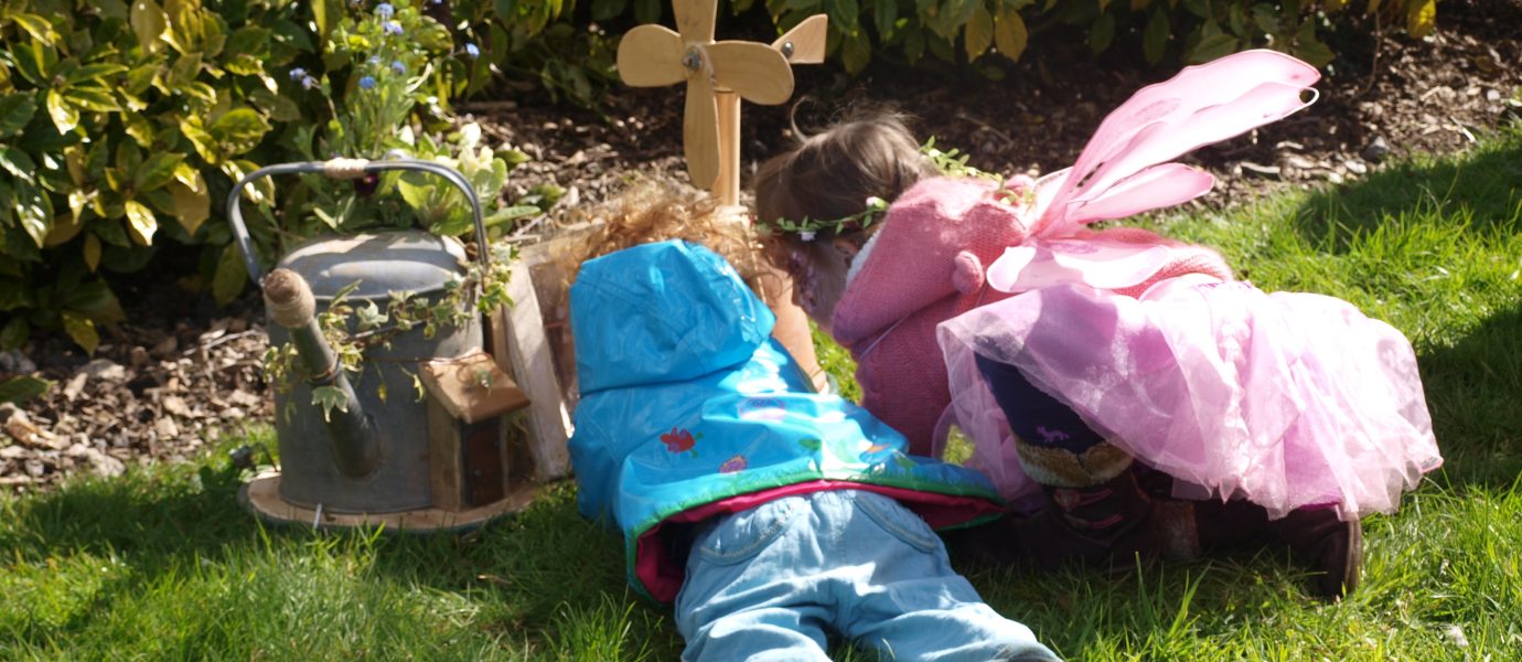 Two young children, one in a blue jacket and the other dressed as a fairy in pink, lie on the grass facing a small wooden cross and watering can, surrounded by green bushes and sunshine.