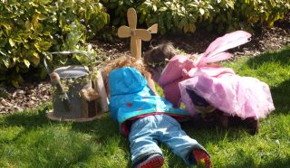 Two young children, one in a blue jacket and the other dressed as a fairy in pink, lie on the grass facing a small wooden cross and watering can, surrounded by green bushes and sunshine.