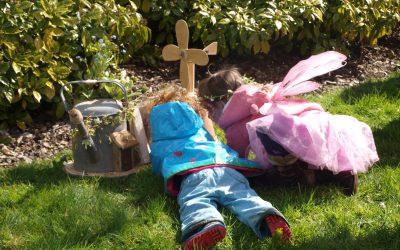 Two young children, one in a blue jacket and the other dressed as a fairy in pink, lie on the grass facing a small wooden cross and watering can, surrounded by green bushes and sunshine.