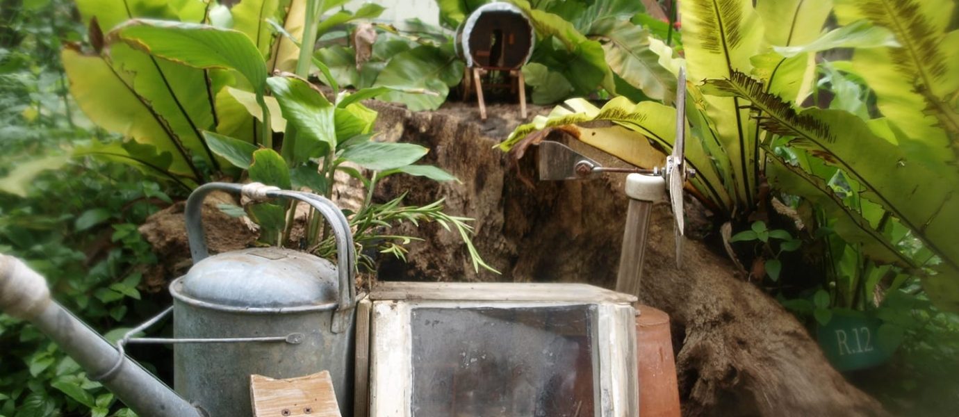 A whimsical garden scene features a miniature house made from a watering can, surrounded by lush green plants, with a small R.I.P. sign on a planter and another tiny house on a log in the background.