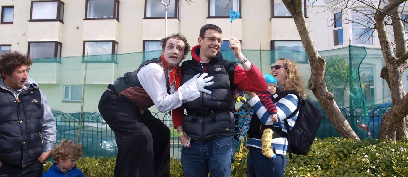 A street performer on stilts poses with a smiling man while a woman holds a small child and waves a blue flag. Other people, including children, stand nearby in front of a building and greenery.
