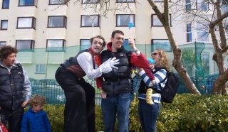 A street performer on stilts poses with a smiling man while a woman holds a small child and waves a blue flag. Other people, including children, stand nearby in front of a building and greenery.