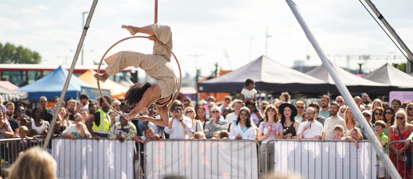 A performer in beige clothing does an acrobatic pose on a suspended hoop at an outdoor festival, while a large crowd watches behind metal barriers and marquees. The sky is partly cloudy.
