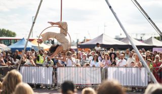 A performer in beige clothing does an acrobatic pose on a suspended hoop at an outdoor festival, while a large crowd watches behind metal barriers and marquees. The sky is partly cloudy.