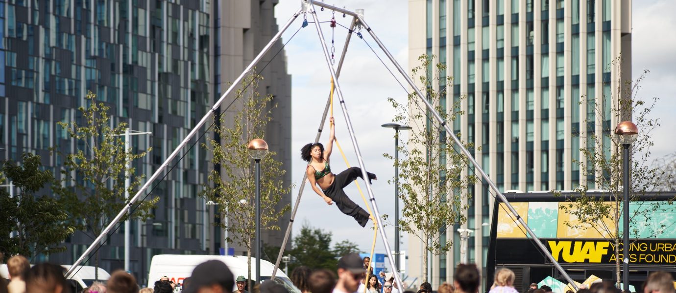 A performer hangs from an aerial hoop on a street stage, surrounded by a large crowd of people at an outdoor urban event with tall modern buildings in the background.