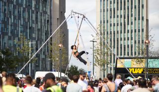A performer hangs on a trapeze in an outdoor urban area, surrounded by a large crowd. Tall modern buildings and trees are visible in the background under a partly cloudy sky.