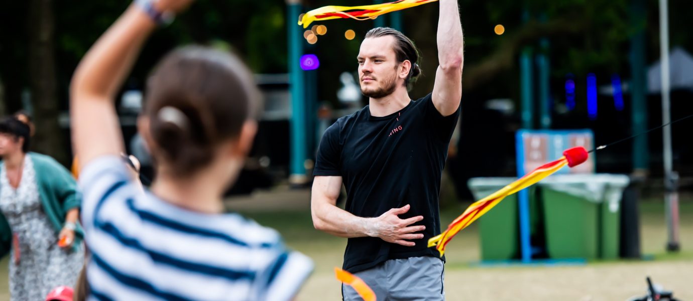 A man in a black shirt skilfully twirls a ribbon outdoors, one arm raised as he performs. A woman in the foreground, back to the camera, also holds a ribbon. People and trees are visible in the background.