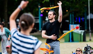 A man in a black shirt skilfully twirls a ribbon outdoors, one arm raised as he performs. A woman in the foreground, back to the camera, also holds a ribbon. People and trees are visible in the background.