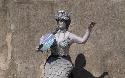 A person dressed as a metallic mermaid poses against a weathered wall, holding a shiny, multicoloured fan. The costume features silver scales, shells, and a decorative headpiece, with silver body paint covering their skin.