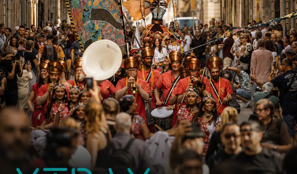 A colourful street parade with performers in elaborate costumes, holding a large decorative banner, moves through a crowded urban street. Text overlays read: XTRAX International Cultural Collaboration & Strategic Support for Outdoor Arts.