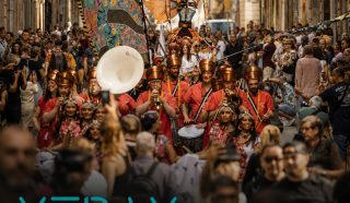 A colourful street parade with performers in elaborate costumes, holding a large decorative banner, moves through a crowded urban street. Text overlays read: XTRAX International Cultural Collaboration & Strategic Support for Outdoor Arts.