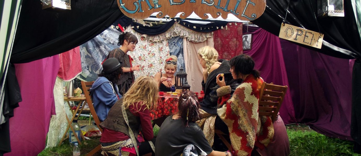 A group of people in colourful costumes sit around a table in a tent decorated with fabric and signs reading “Café Petite” and “Open,” creating a whimsical, cosy atmosphere at an outdoor event.