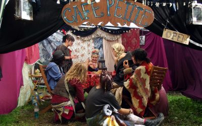 A group of people in colourful, bohemian clothing sit around a table in a tent decorated with fabrics and a sign reading Cafe Petite. They appear to be engaged in conversation or a creative activity.