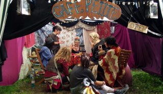 A group of people in colourful costumes sit around a table in a tent decorated with fabric and signs reading “Café Petite” and “Open,” creating a whimsical, cosy atmosphere at an outdoor event.